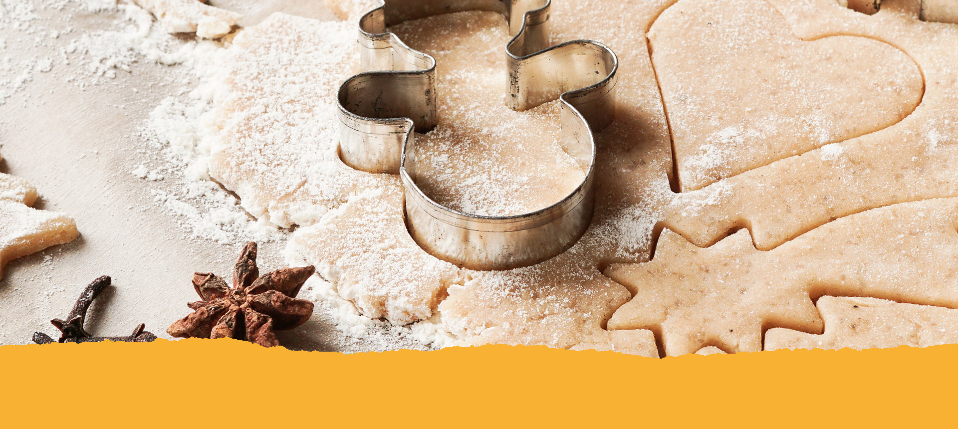 Ausgerollter Plätzchenteig mit ausgestanzten Herzchen- und Sternmotiven. Auf dem Teig liegt ein Schneemann-Keksförmchen und davor ein Sternanis und einige Nelken. Text: Unsere Weihnachtsgewürze