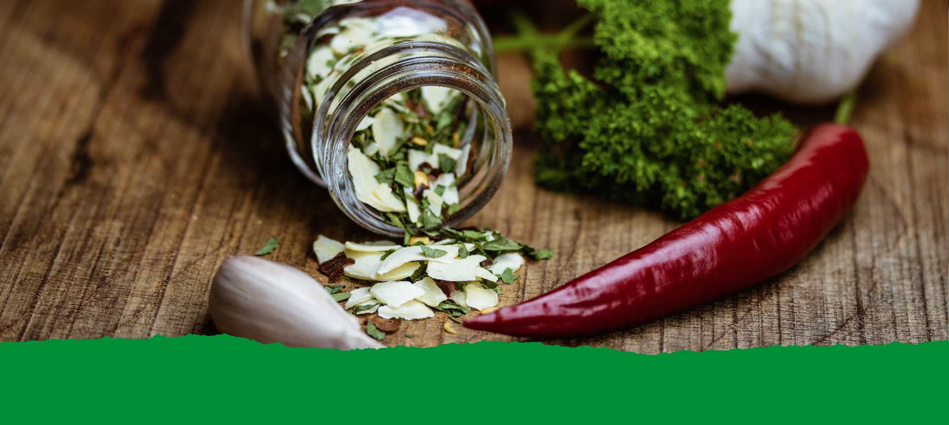 Petersilie, Knoblauch, Chili und ein Glas mit getrockneten Gewürzen auf dunklem Holz. Text: Unsere Kräuter und Gewürze