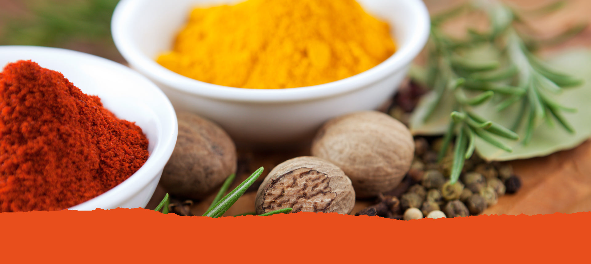 Ein Schälchen mit rotem Gewürz- und eines mit gelbem Gewürzpulver, davor einige Muskatnüsse, Pefferkörner und einige grüne Kräuter. Text: Unsere Gewürzmischungen