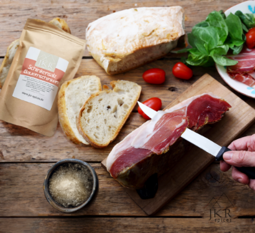 Hand schneidet Schinken auf Holzbrett, daneben Brot, Tomaten, Basilikum und JKR Schinkensalz Bauernschinken Packung