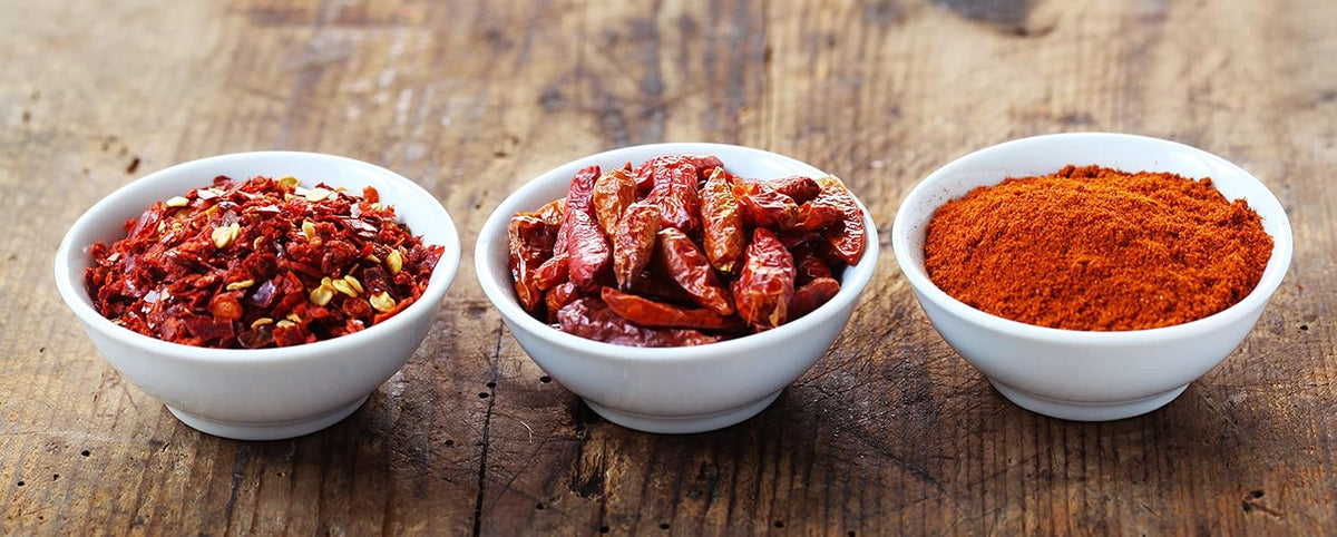 Chili und Peperoncino Gewürze als Zutatenmotiv, Chiliflocken, getrocknete Schoten und Chilipulver in Schalen auf Holz