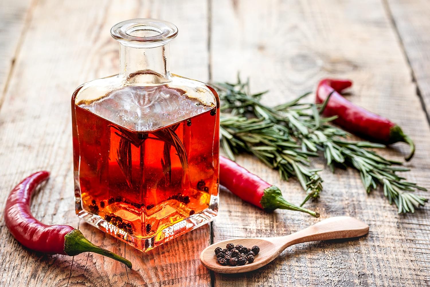 Serviervorschlag Peperoncino Öl, Glasflasche mit Chiliöl, rote Chilischoten, Kräuter und Pfefferkörner auf Holzuntergrund