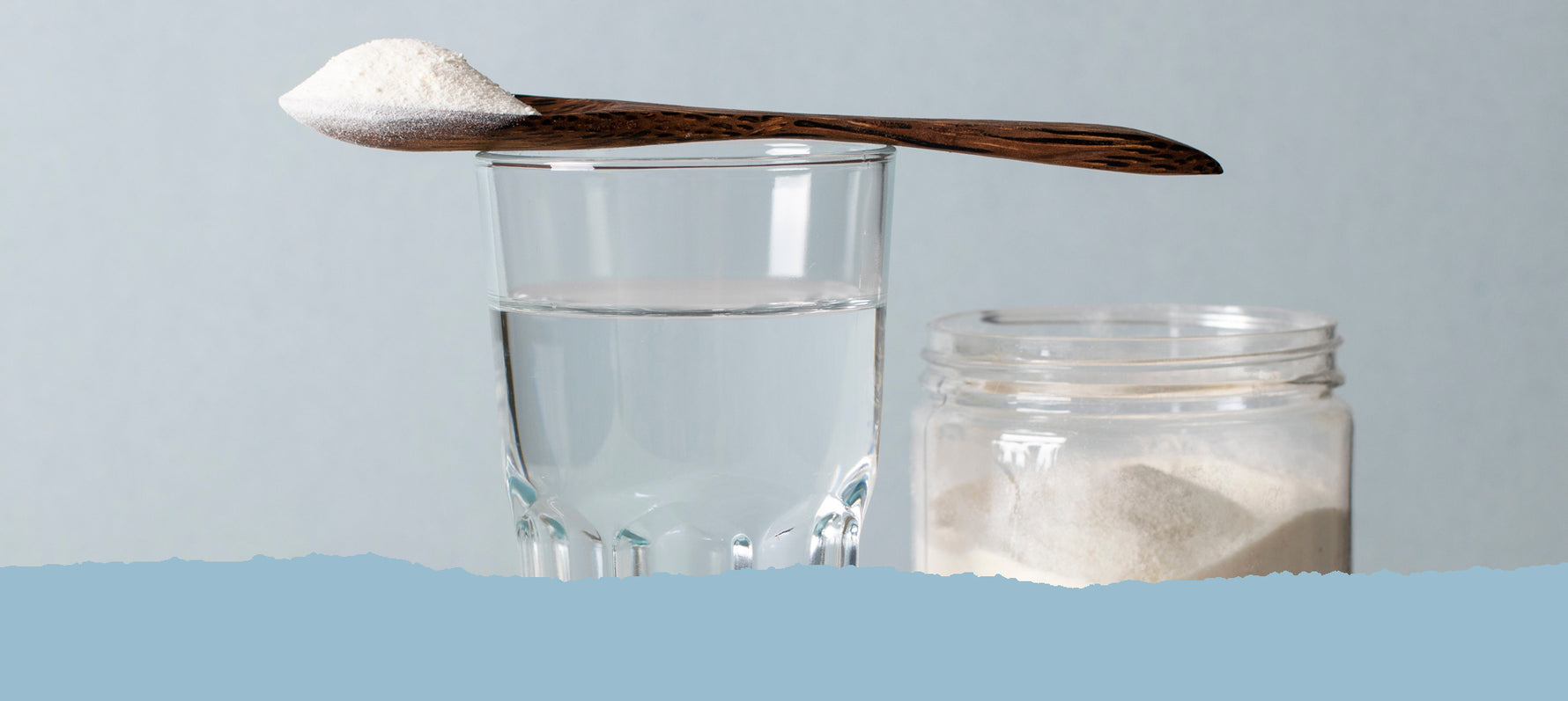 Ein Glas Wasser und ein Glas mit weißem Pulver vor hellblauem Hintergrund. Auf dem Rand des Trinkglases liegt ein Holzlöffel mit weißem Pulver. Text: Unsere Supplements / Minerals