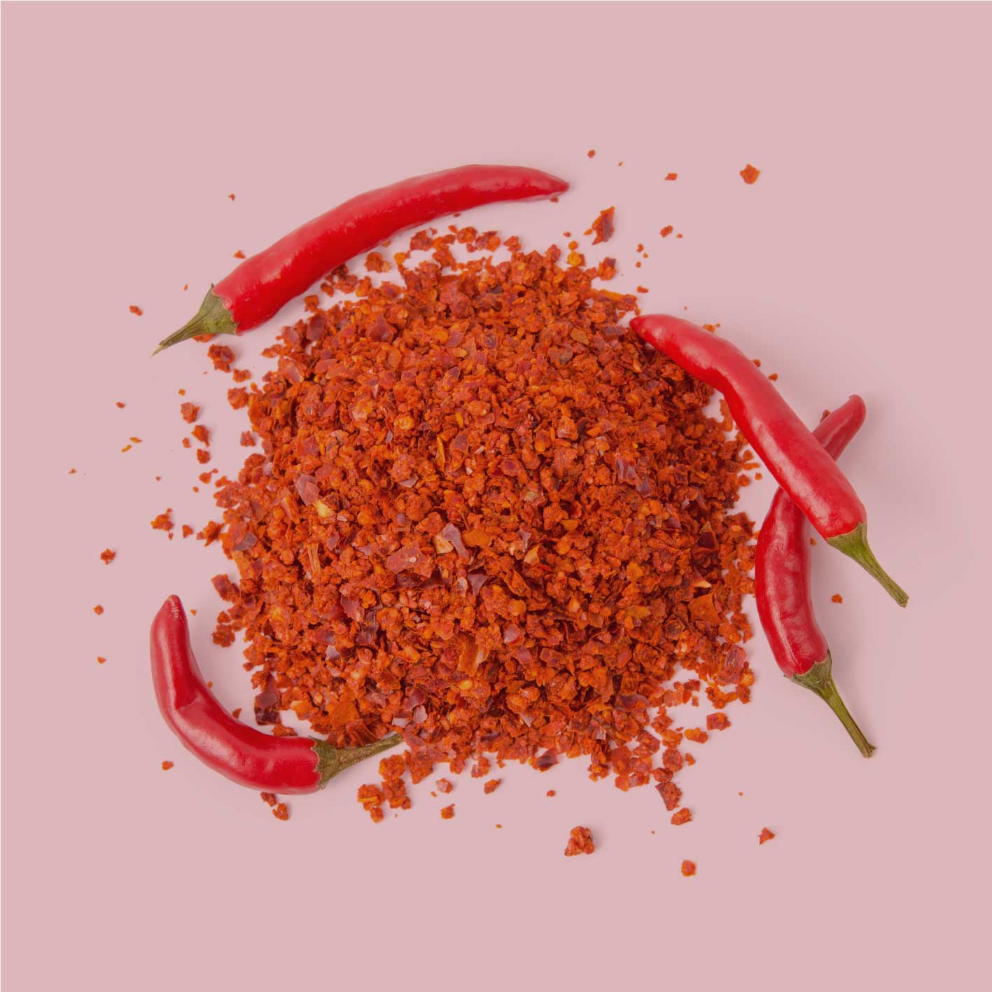Rote Chiliflocken mit frischen Chili­schoten auf rosa Hintergrund – dekorative Produktdarstellung